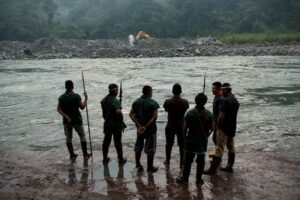 Miembros de la comunidad indígena Cofán de Sinangoe caminan a orillas del río Aguarico, mientras al fondo se ve una retroexcavadora removiendo tierra. Foto: Jerónimo Zuñiga/Amazon Frontlines.